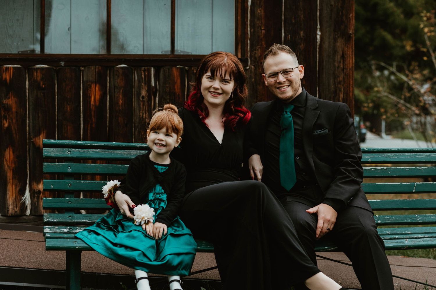A family sits together on a green bench against a rustic wooden building backdrop.