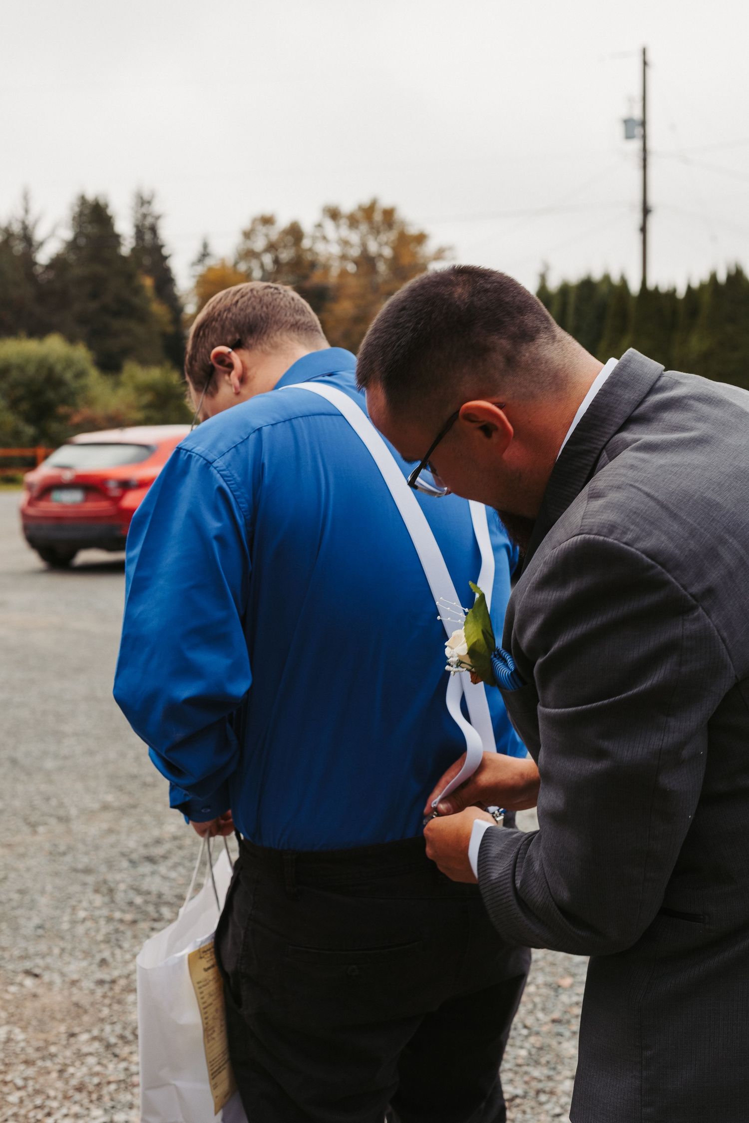 Two people stand together in formal attire while adjusting clothing in an outdoor parking area.