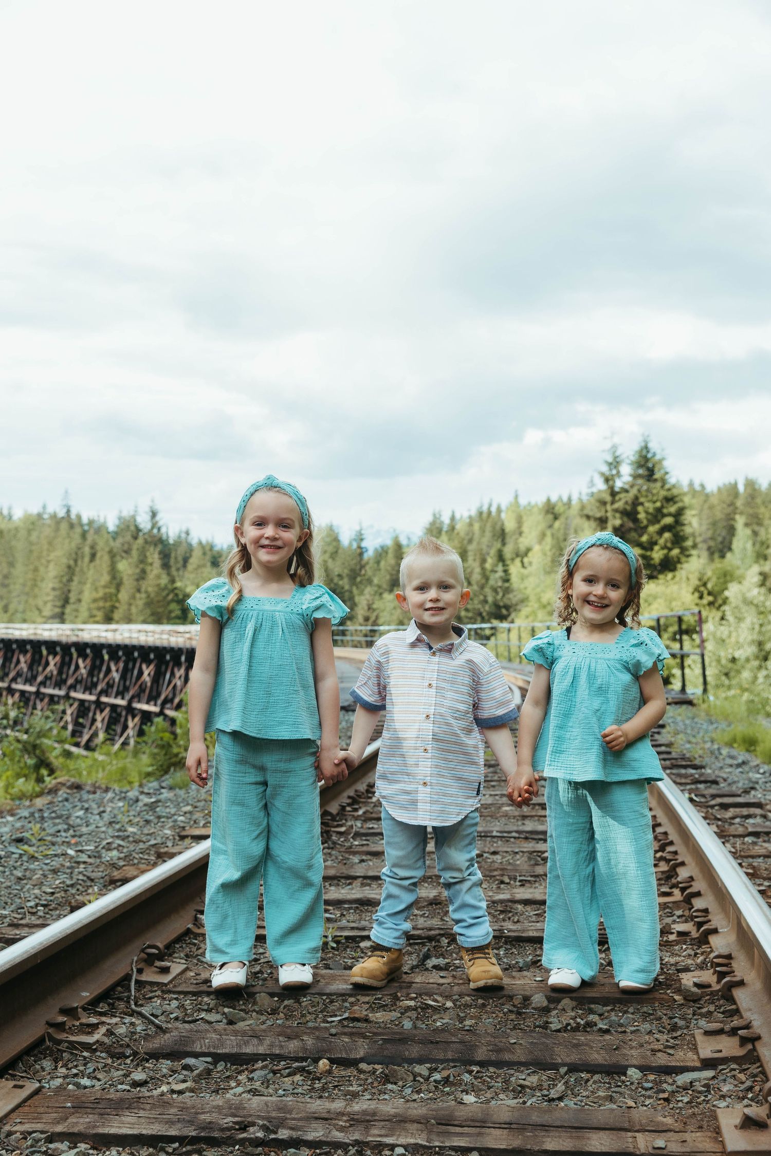 Three small children in coordinating turquoise and white outfits stand on wooden railroad tracks.