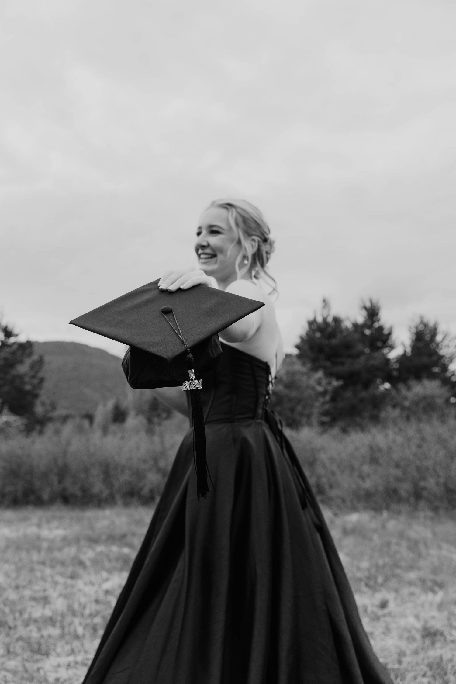 Graduate in a black formal gown poses joyfully in an outdoor field with mountains in the background.