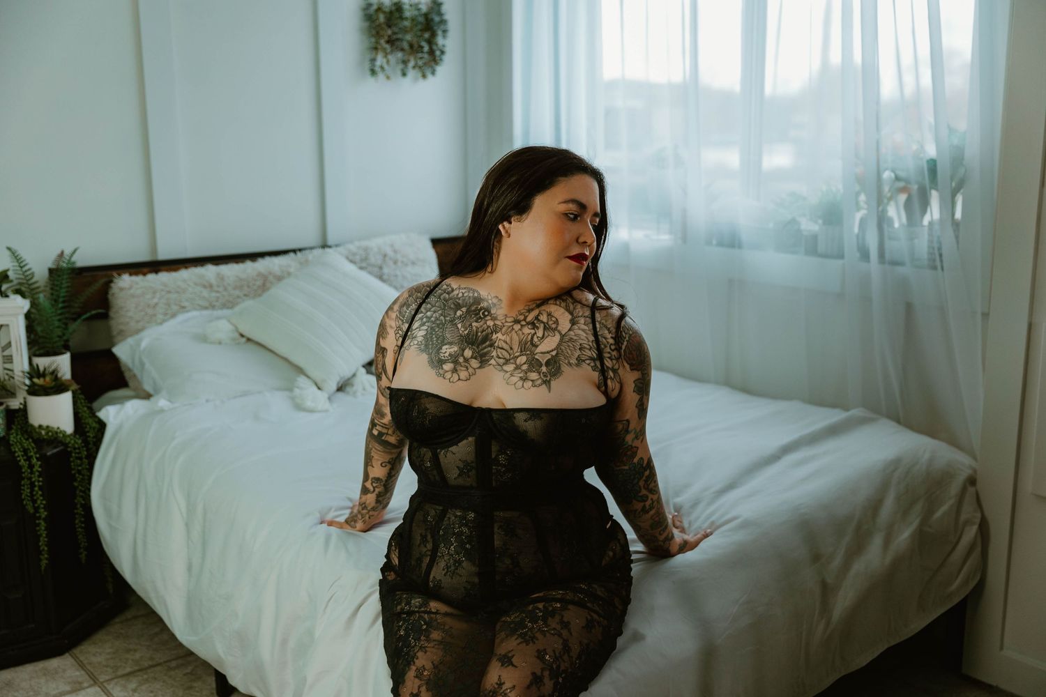 Person in black lace lingerie posing on white bed with plants in background.