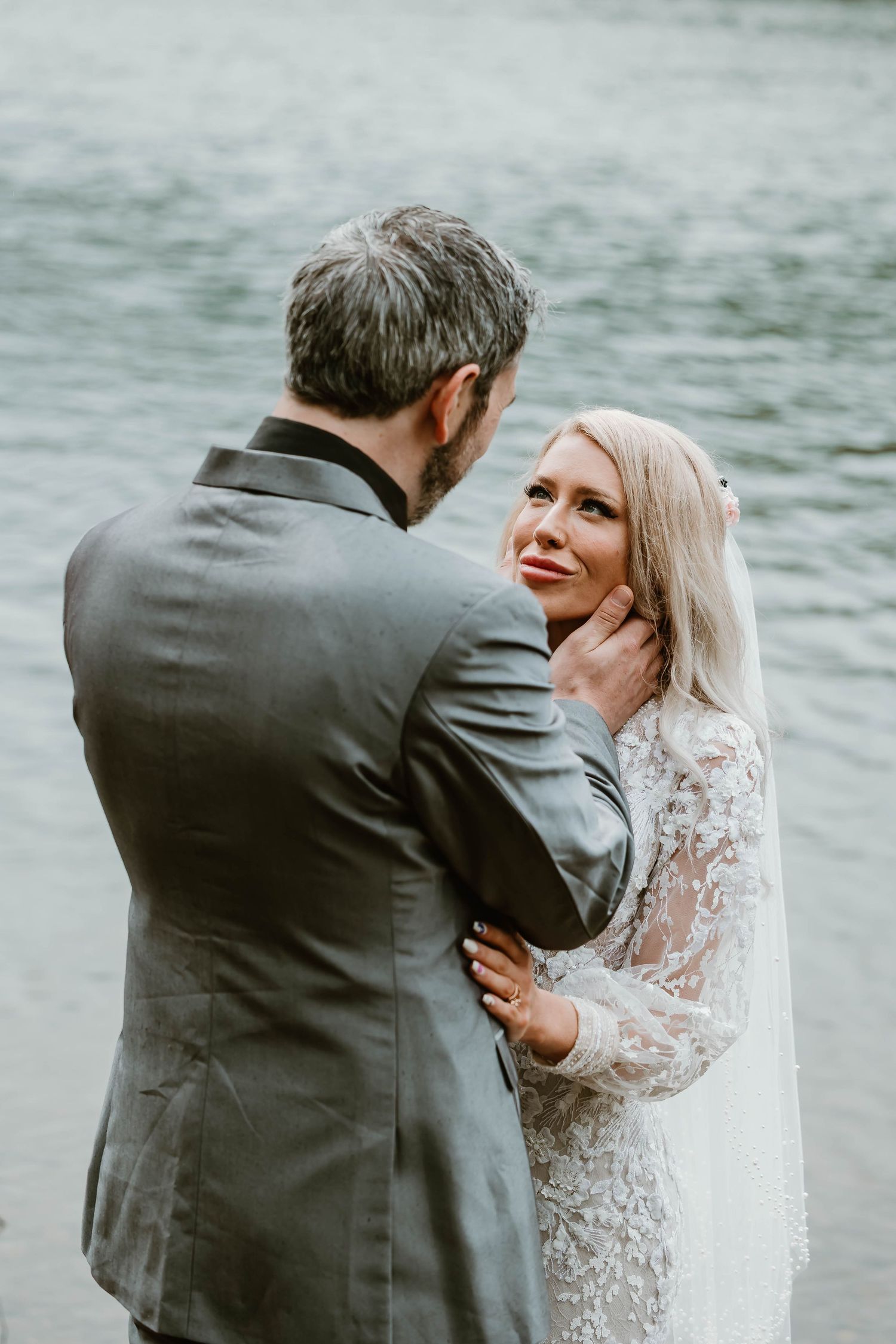 A tender wedding moment captured between a couple in a grey suit and lace gown by the water's edge.