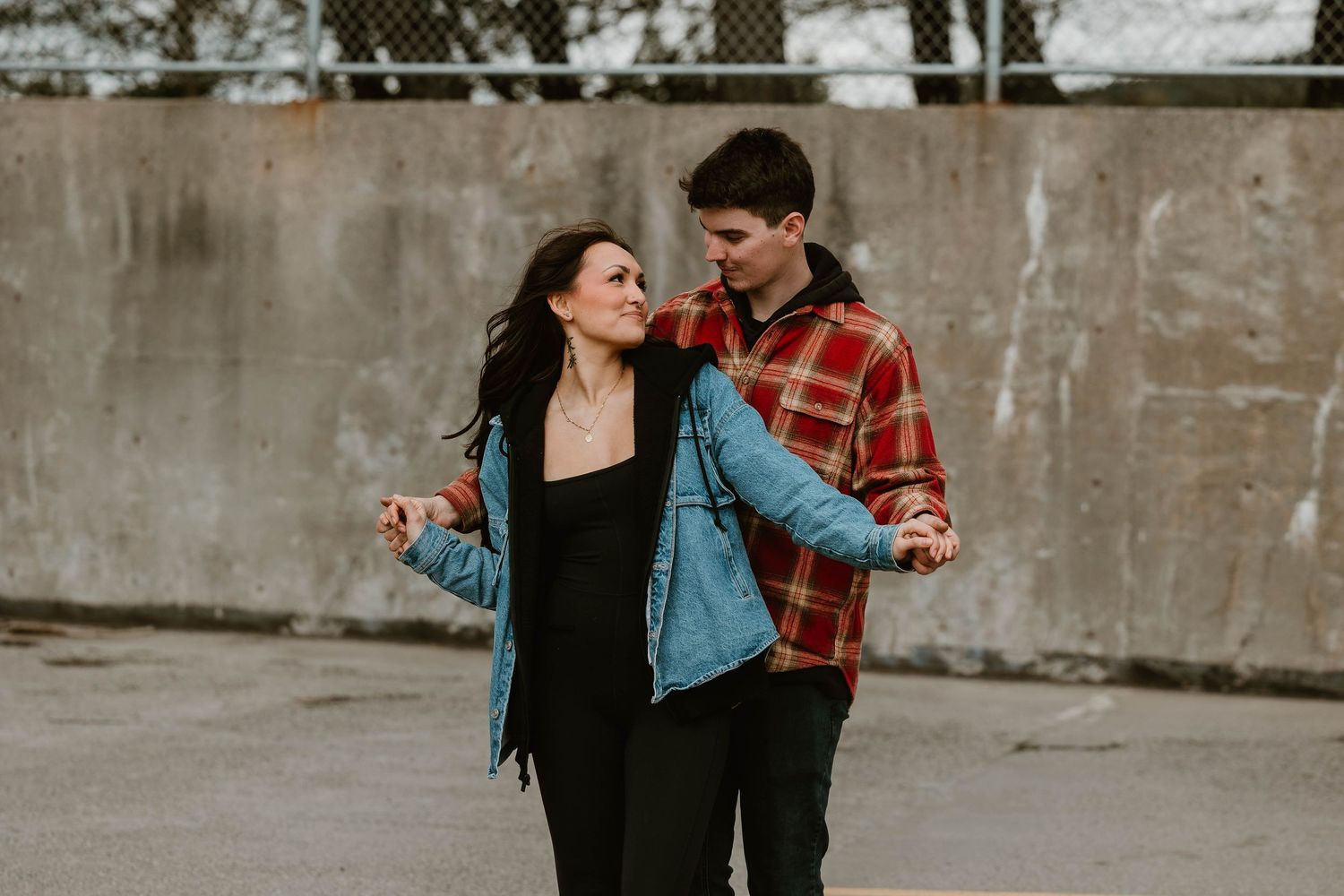 A young playful couple dancing together on a city sidewalk wearing casual denim and plaid clothing.