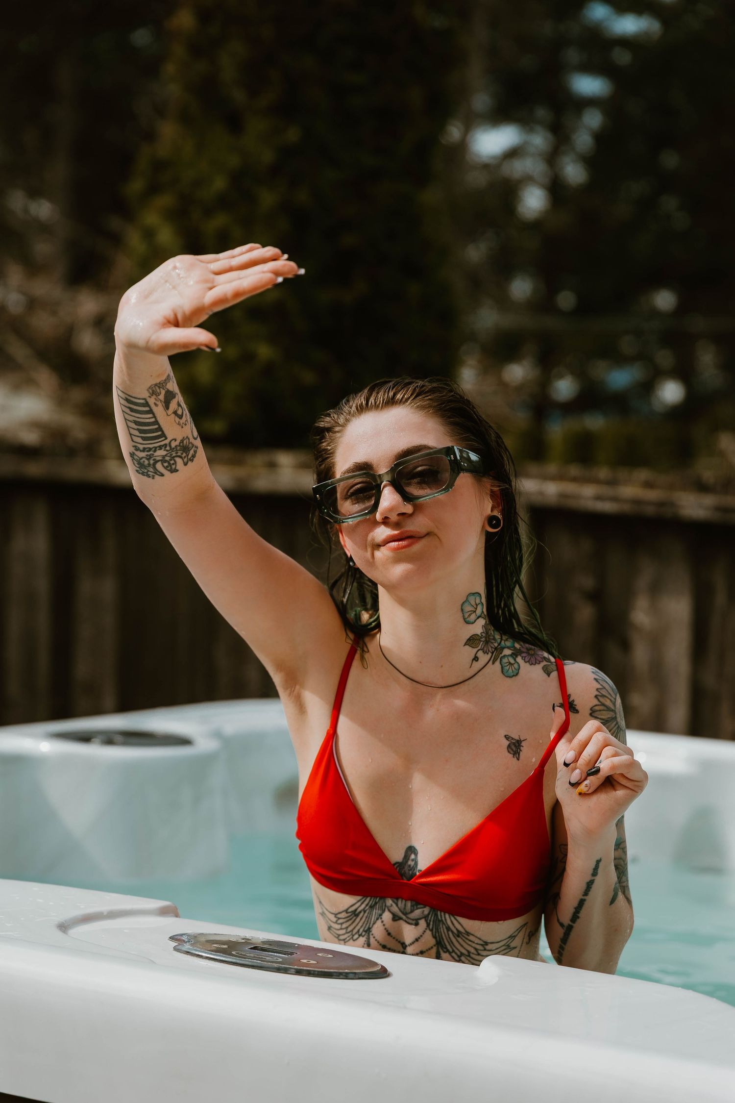 Woman in red swimwear relaxing in outdoor hot tub on sunny day, wearing sunglasses and showing tattoos.