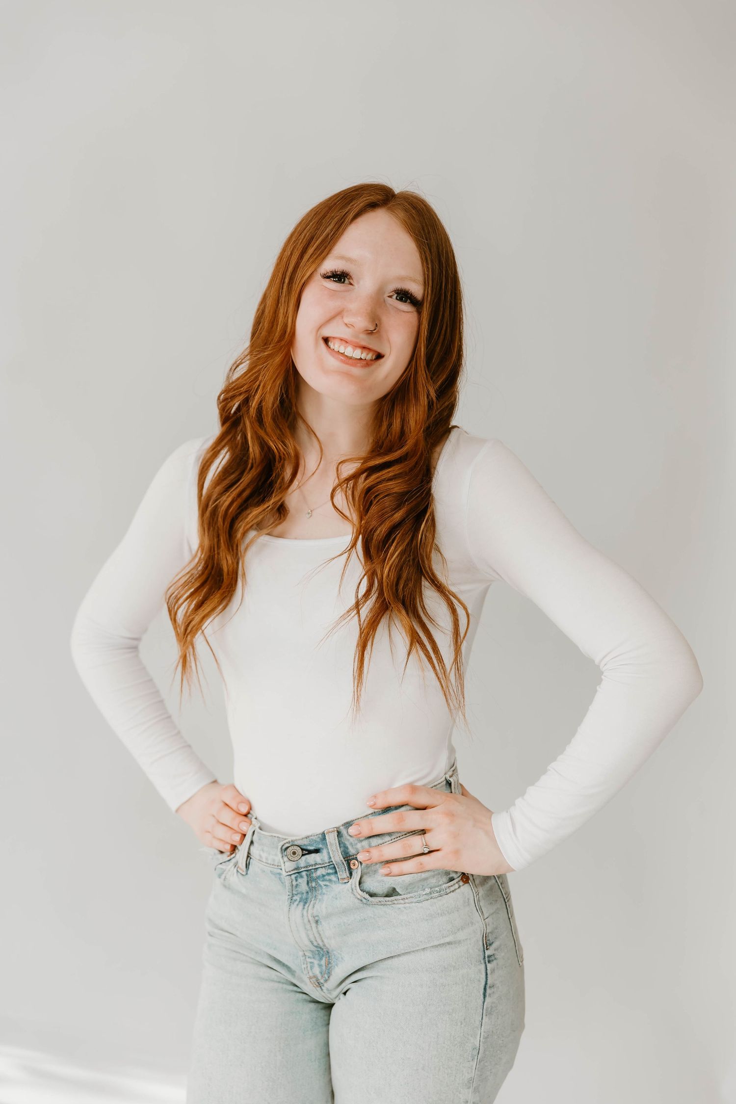 Natural light portrait of a person with long red hair wearing a white long-sleeve top and light wash jeans against neutral backdrop.