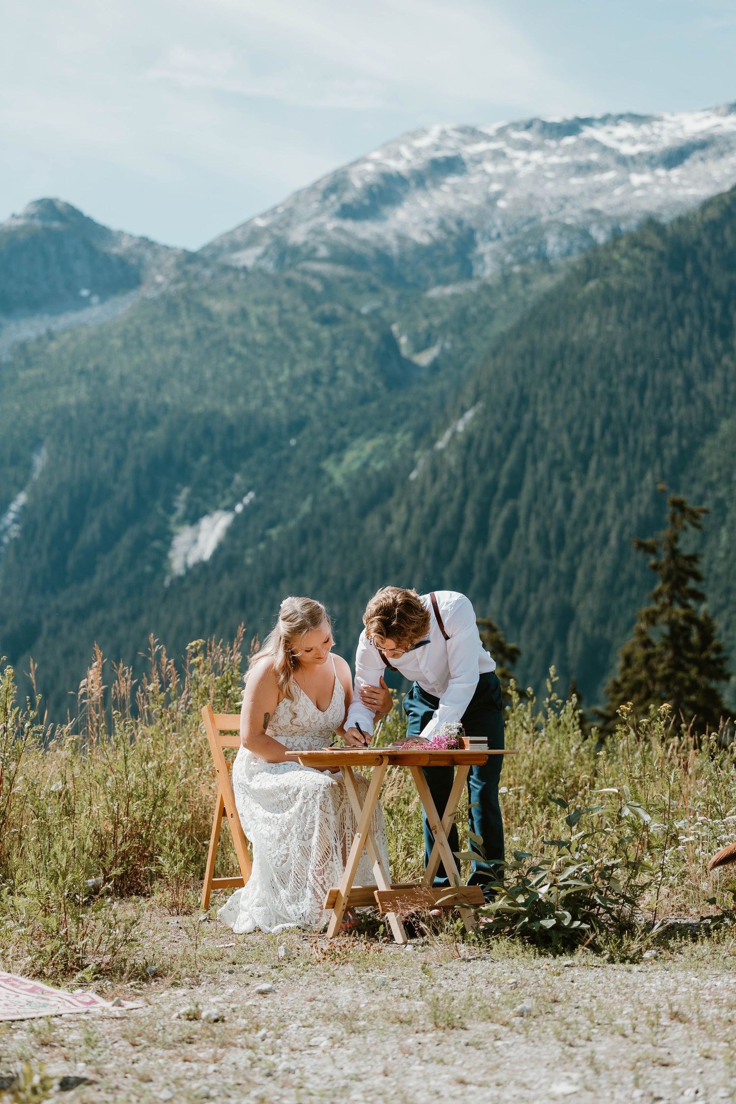 A couple signs their marriage license on a table outdoors with snow-capped mountains in the distance.