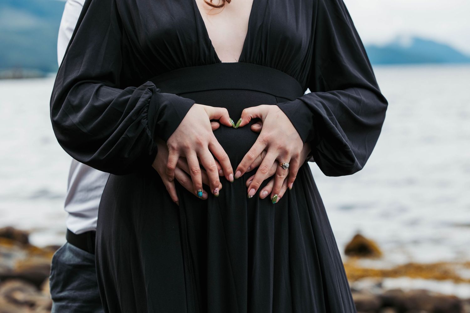 Hands forming a heart shape against a black dress with water and mountains in the background.