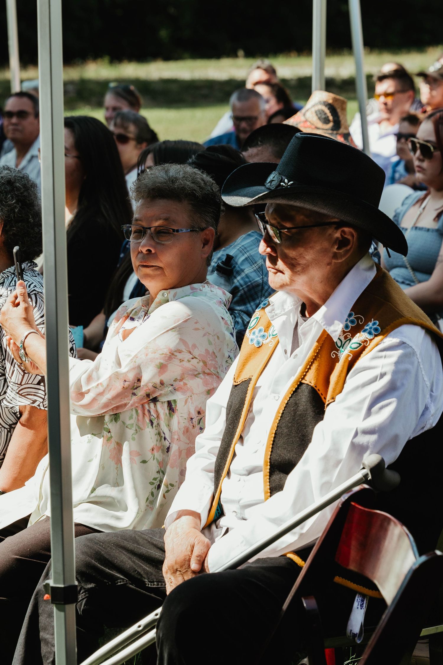 Wedding guests in formal attire seated under a white canopy during an outdoor ceremony.