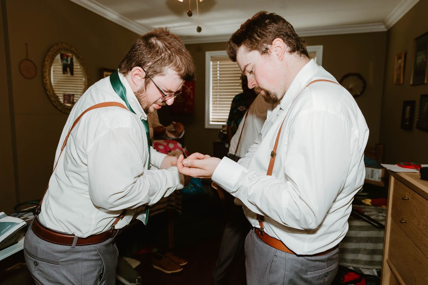 Two groomsmen in suspenders and bowties help each other with final preparations.