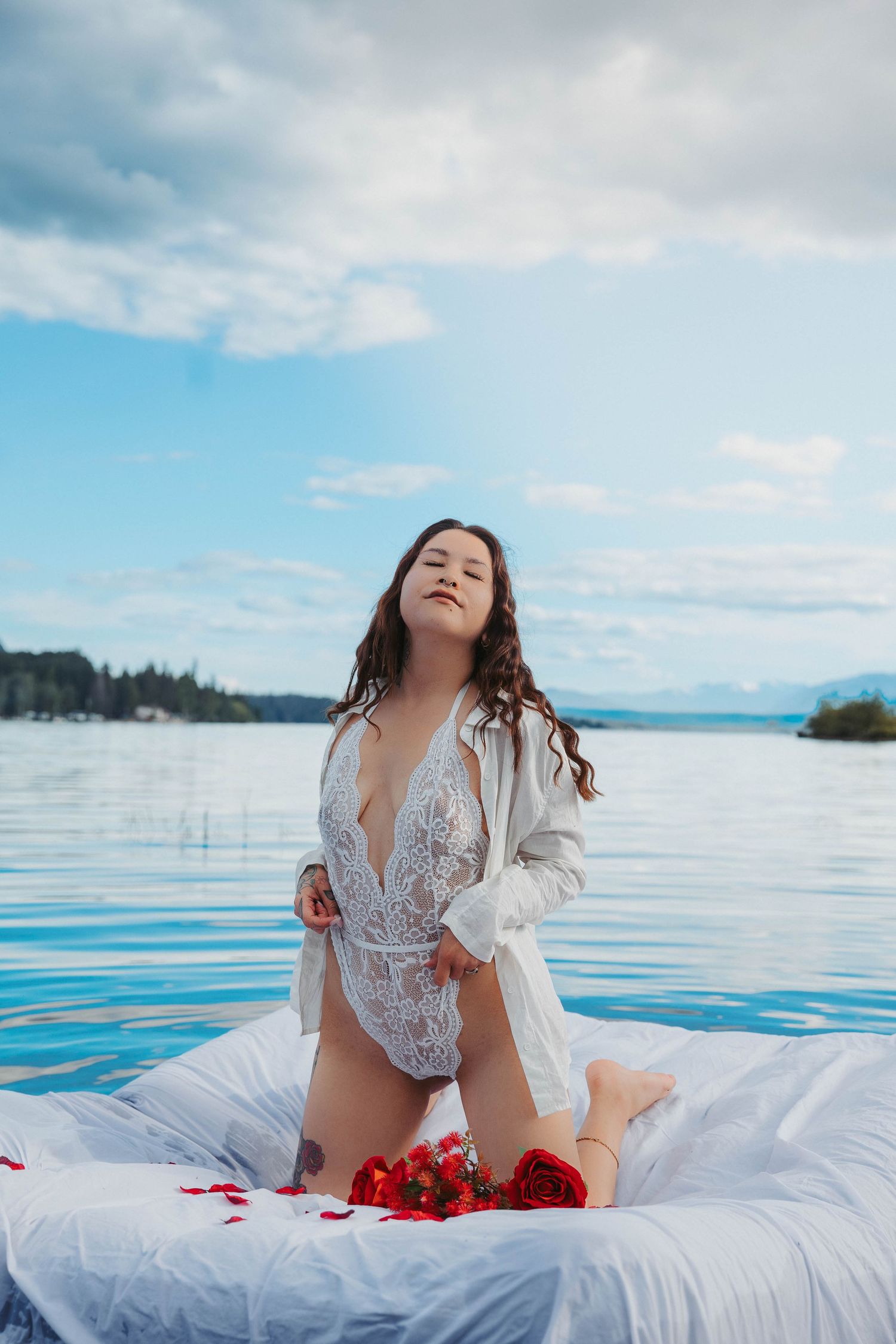 Person in white lace bodysuit poses on white bedding on water with blue sky backdrop.