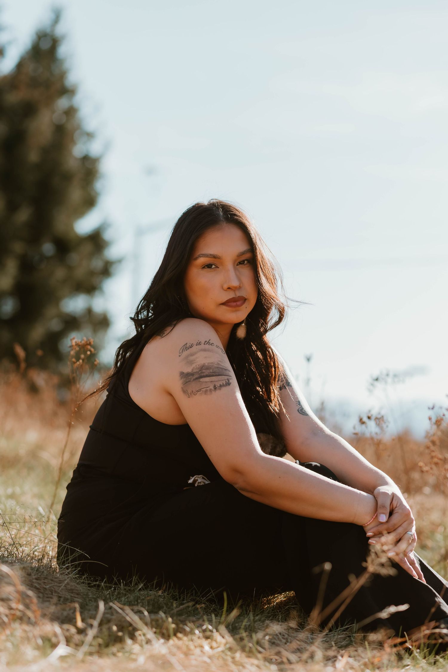 Person in a black dress poses thoughtfully on grass during golden hour in an artistic outdoor portrait series.