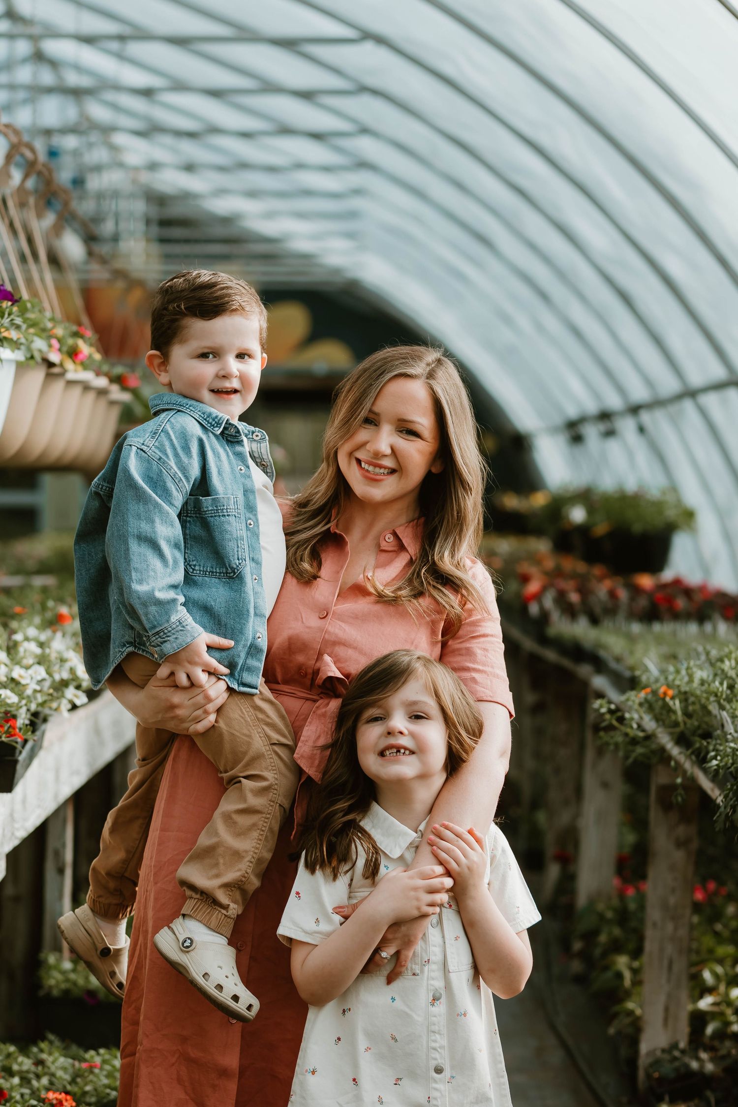 Family poses together inside a greenhouse with hanging plants and flowers in a warm natural setting.