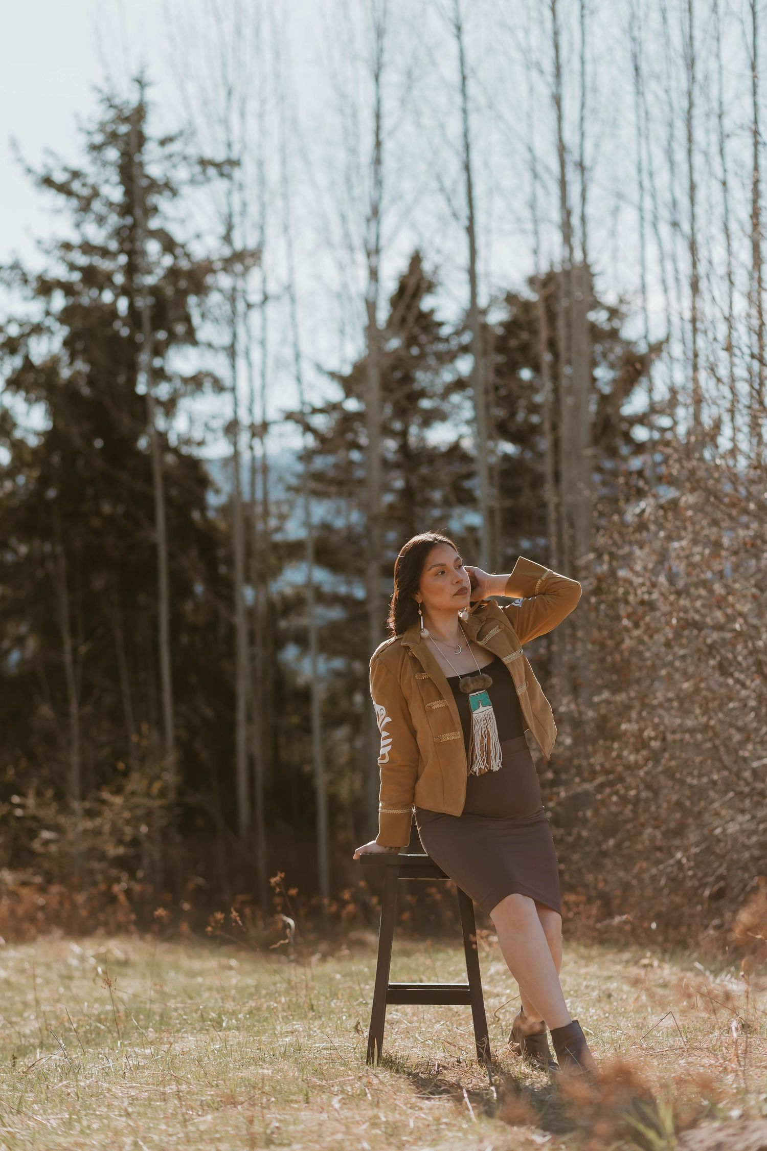 A person in a brown jacket and scarf sits on a wooden stool outdoors against a backdrop of evergreen trees on an autumn day.
