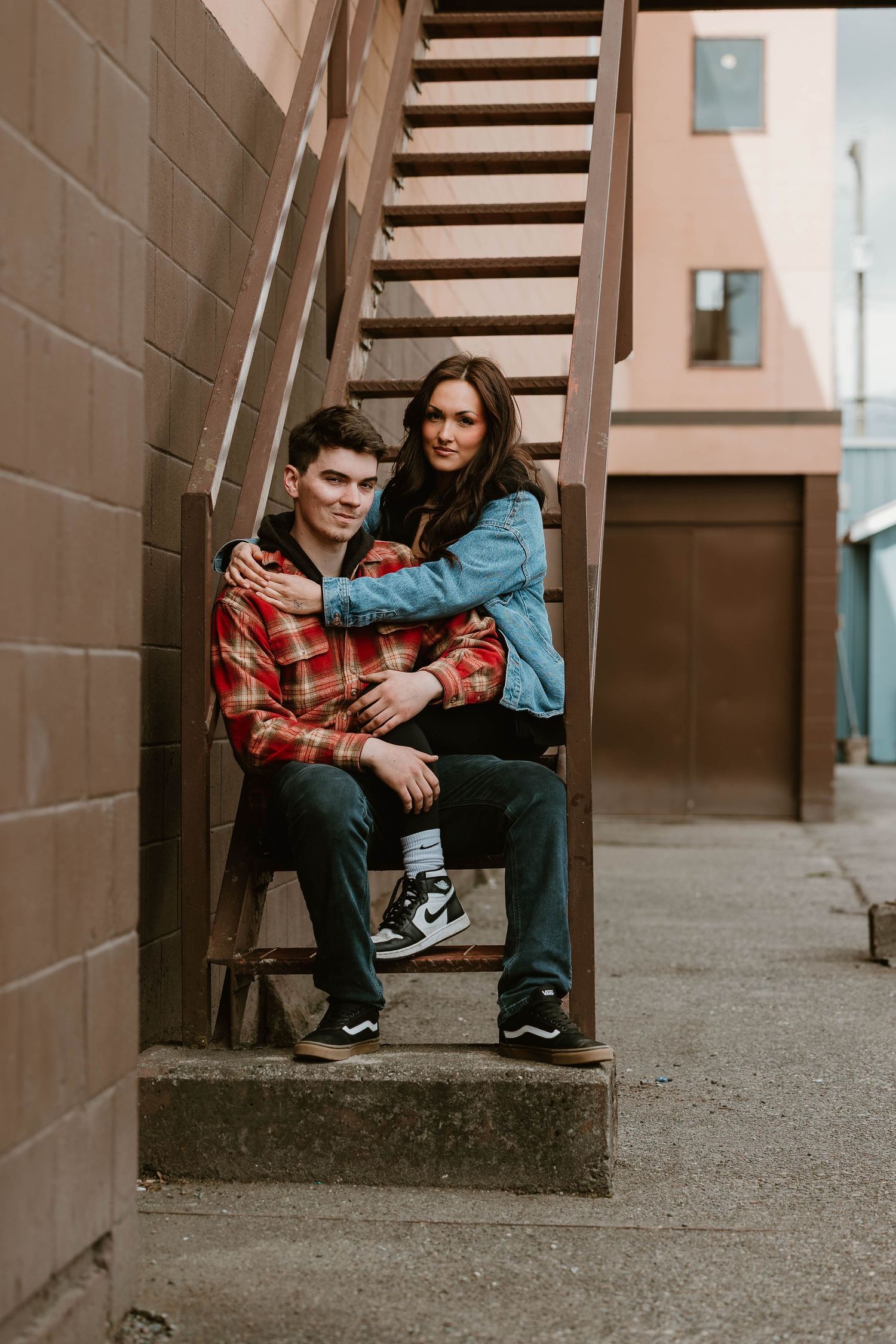 Couple sitting together on outdoor concrete steps against brick wall in urban setting wearing casual denim and plaid.