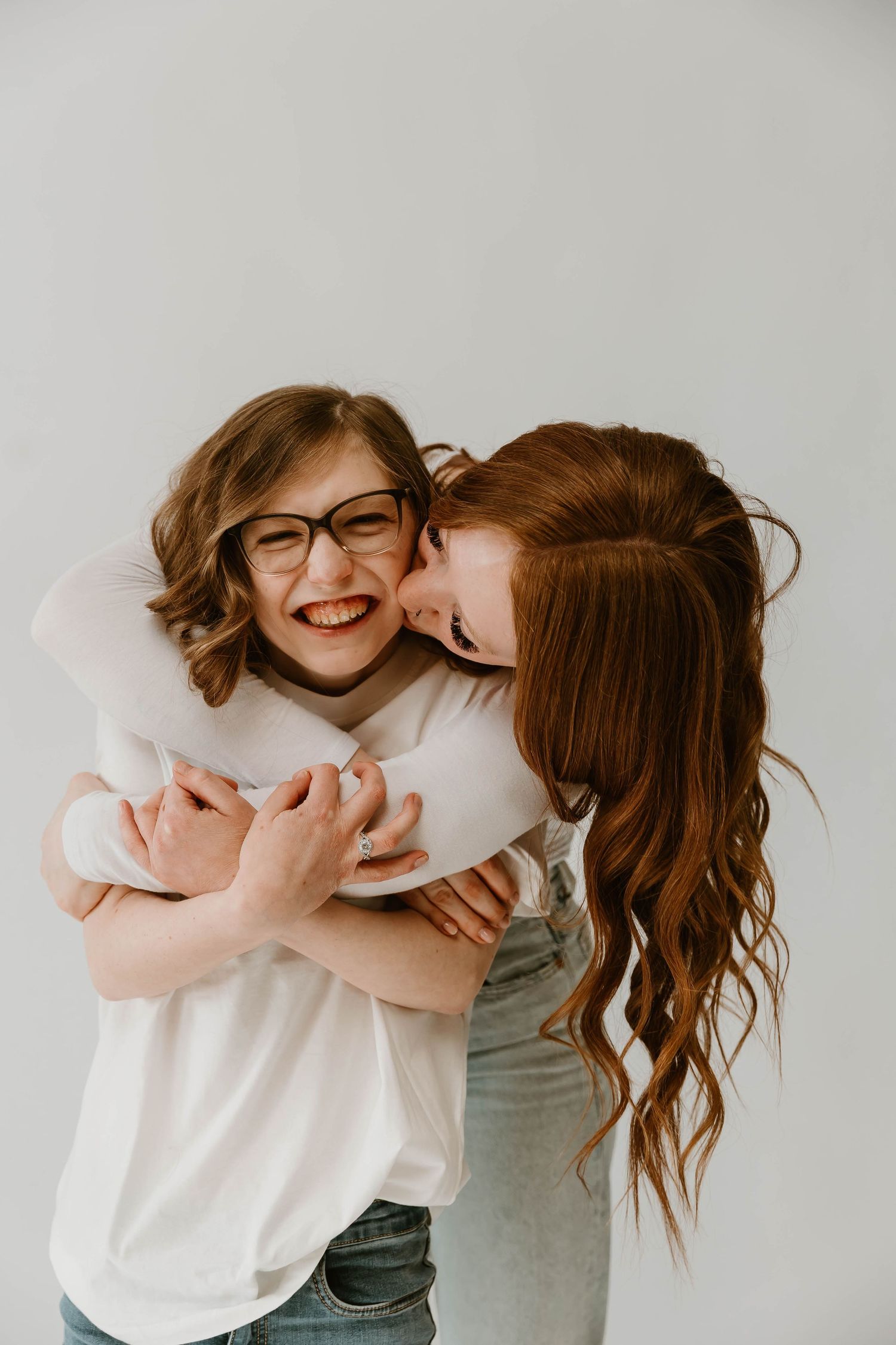 Two friends share a joyful moment of laughter and affection while wearing casual clothing against a light background.