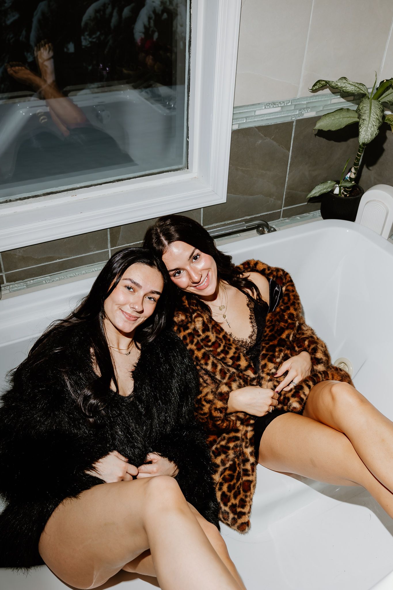 Two people in casual attire relax together on a white surface near windows with plants visible outside.