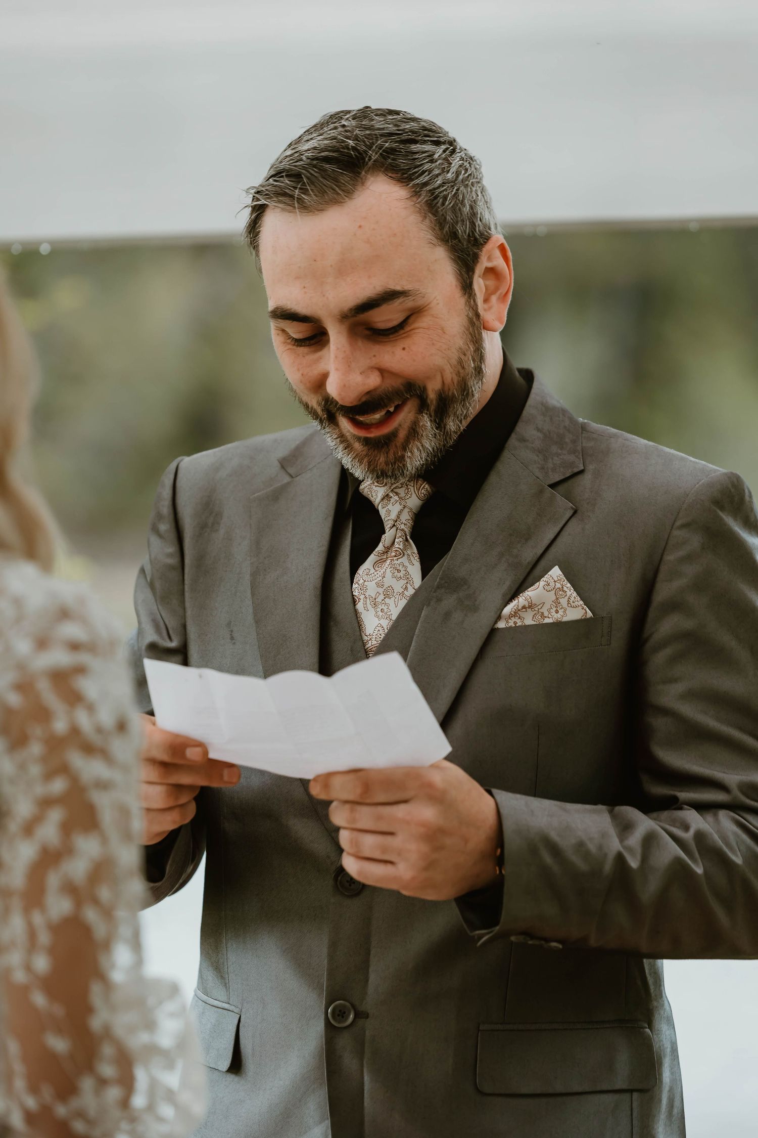A groom in a brown suit and patterned tie smiles while reading from a paper during an outdoor wedding ceremony.