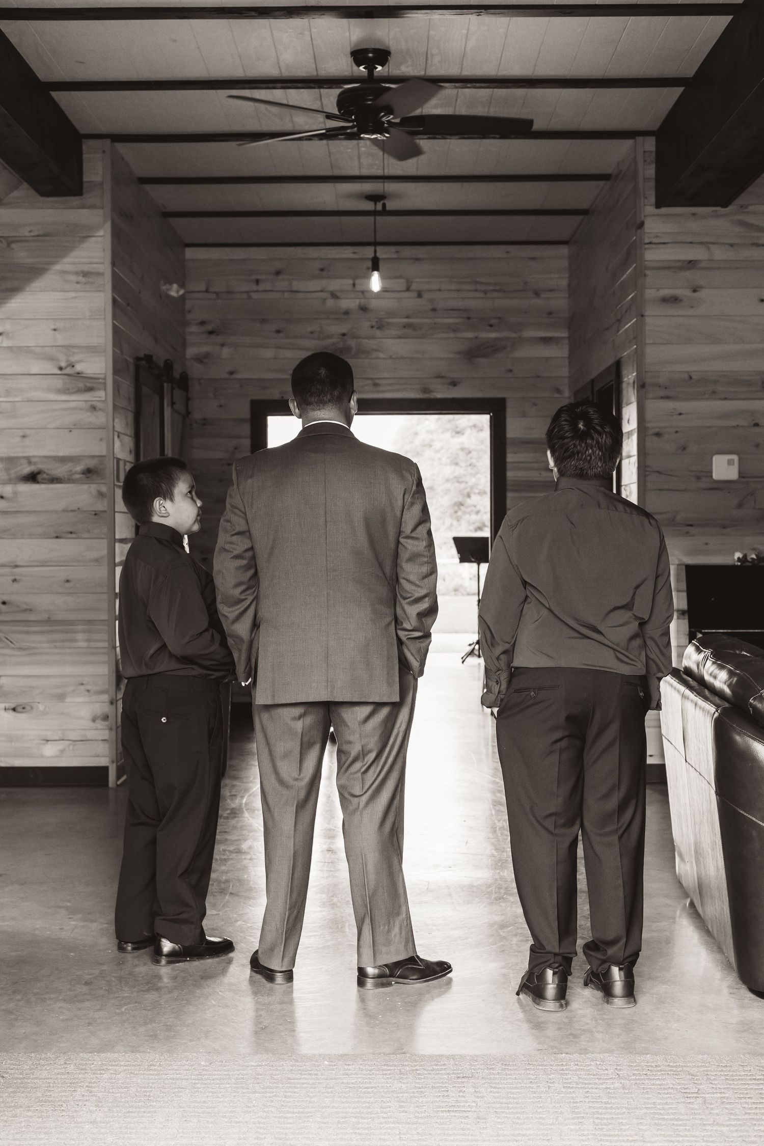 Three people in formal attire stand looking out a window in a wood-paneled room.