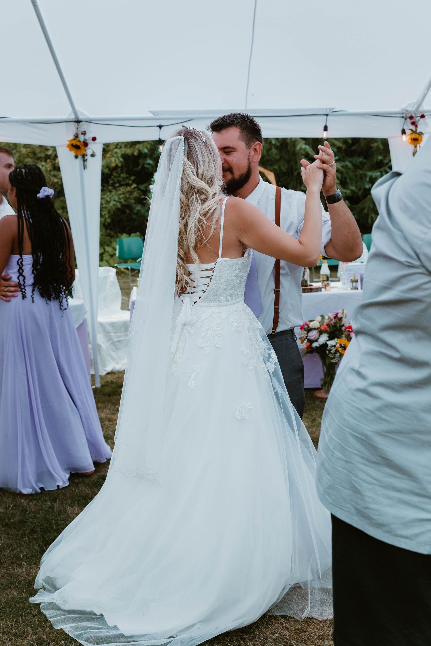 A bride in a flowing white dress dances with someone wearing suspenders under a white tent at an outdoor wedding reception.