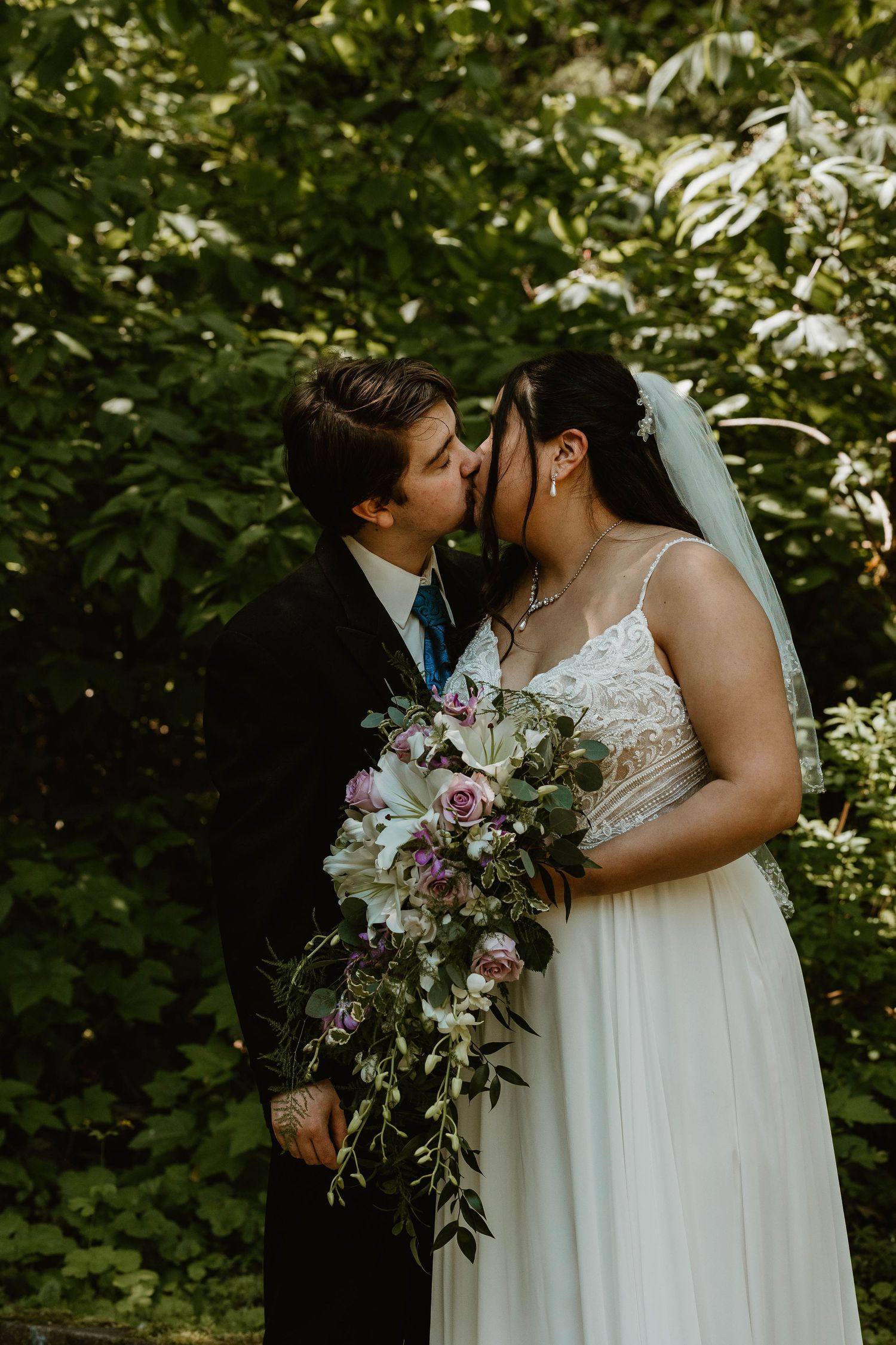 Romantic wedding photo of couple embracing among green foliage with a floral bouquet.