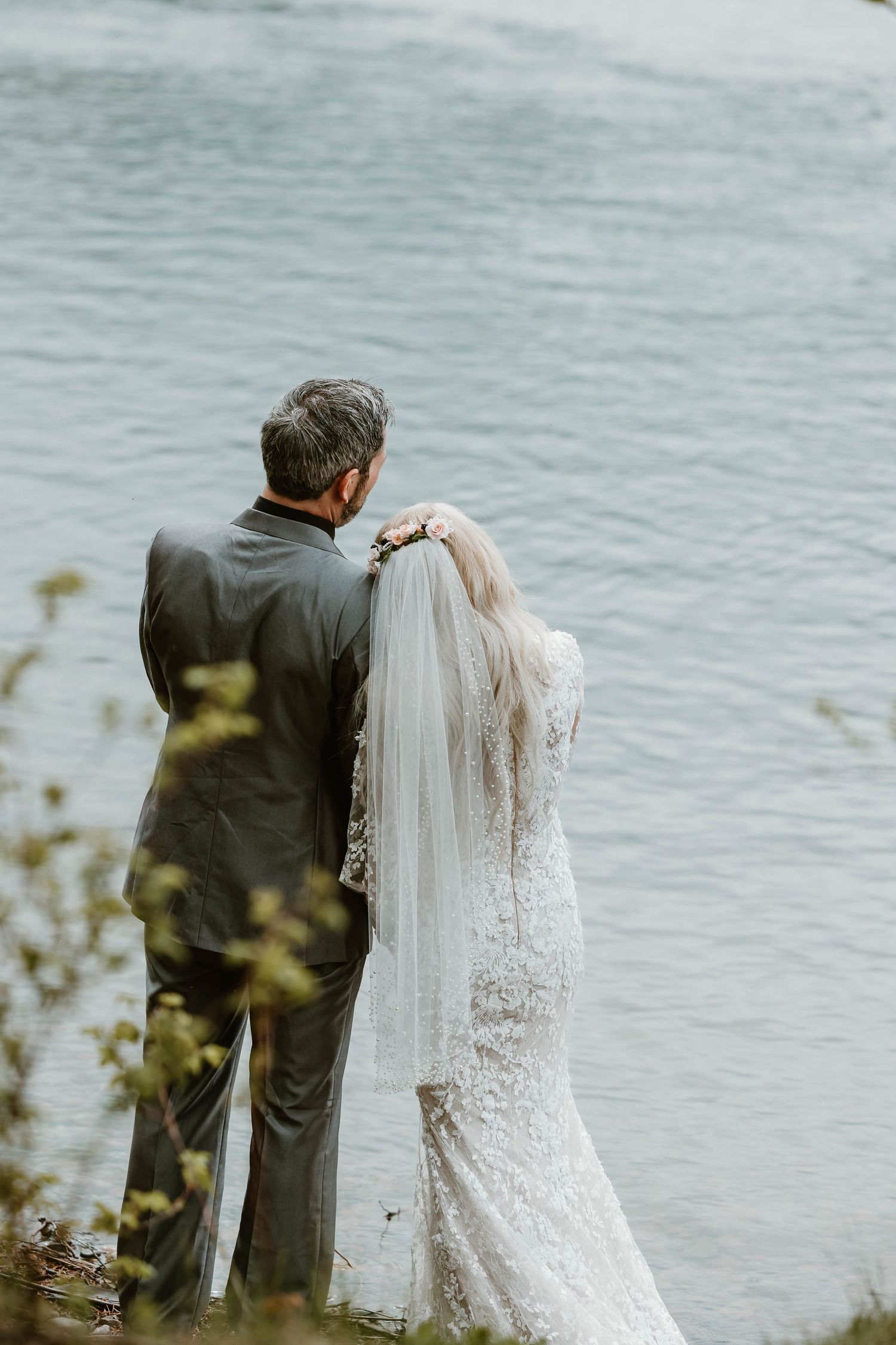 A bride and groom share an intimate moment while standing together near a serene waterfront on their wedding day.