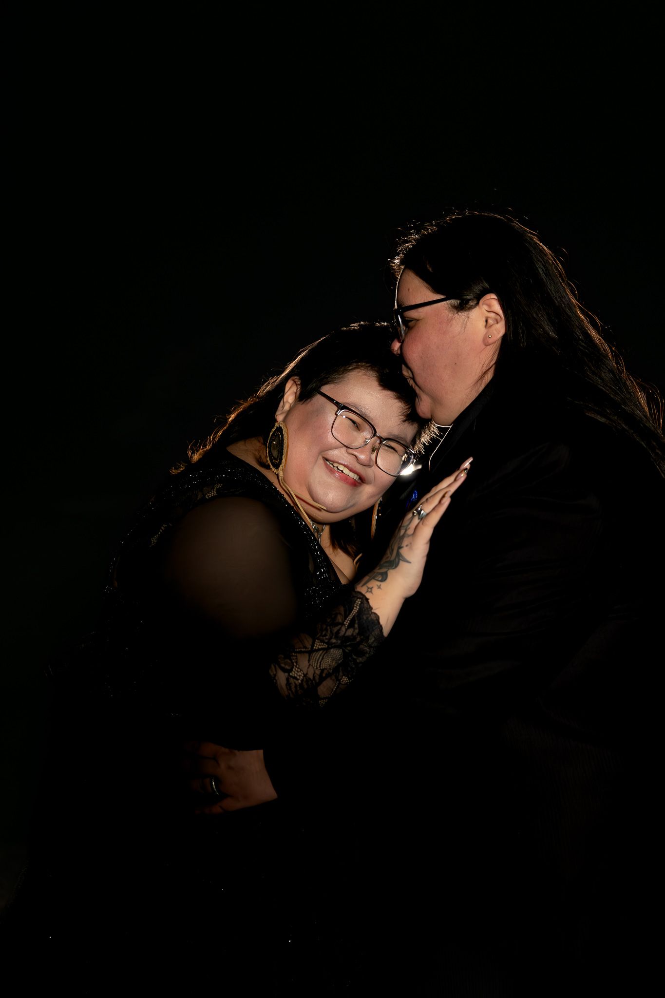 A romantic couple embraces in a dark studio setting with dramatic lighting and shadows.