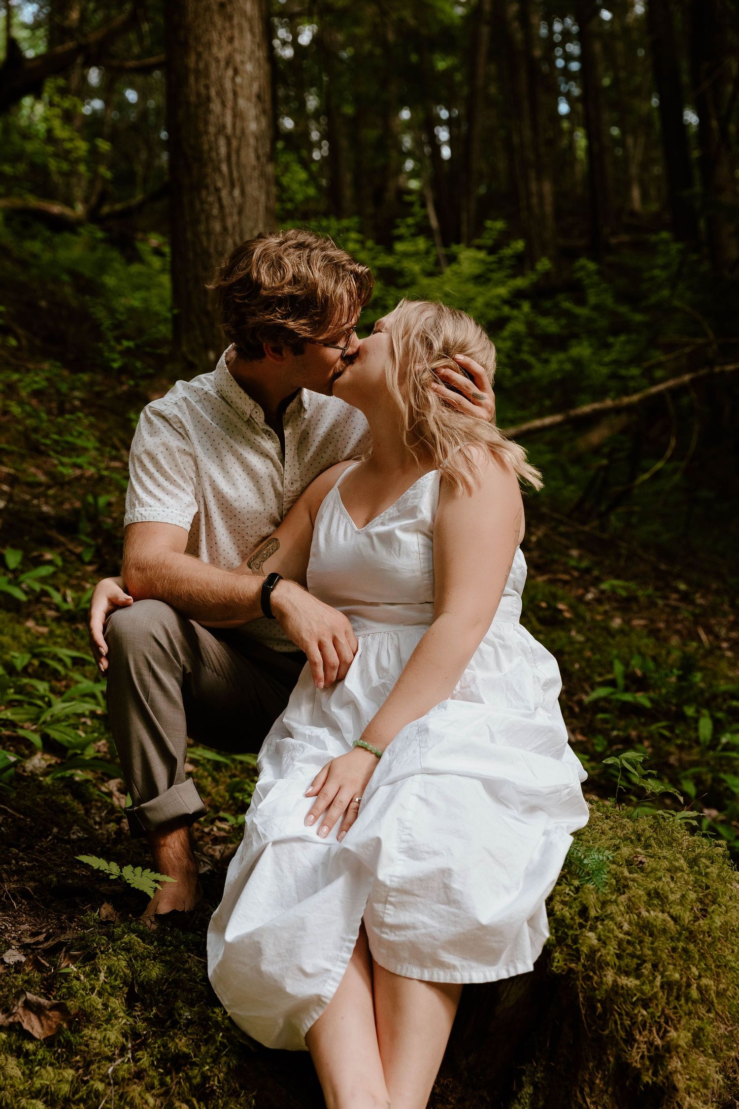 Romantic couple in white clothing share an intimate moment in a mossy forest setting.