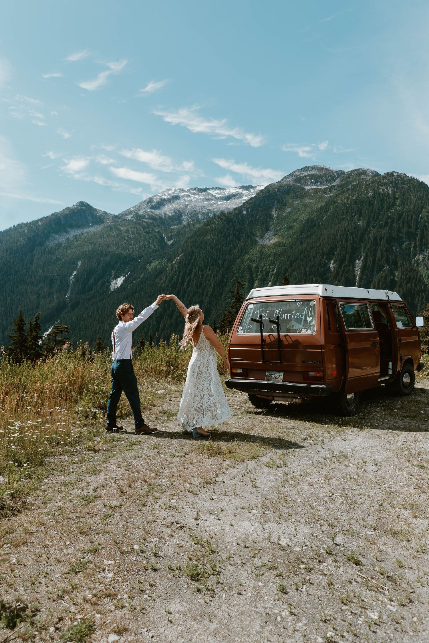 A vintage orange van parked in front of snow-capped mountains with a couple dancing nearby.