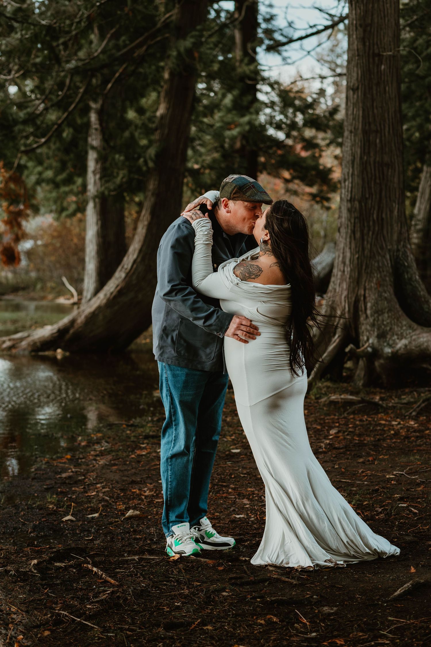 A romantic moment between a couple in white dress and casual wear among tall trees by the water.