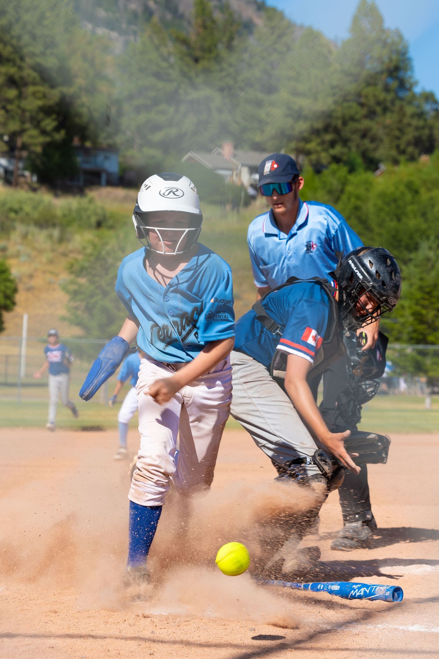 Youth baseball player in light blue uniform sliding into home plate as the ball rolls nearby.