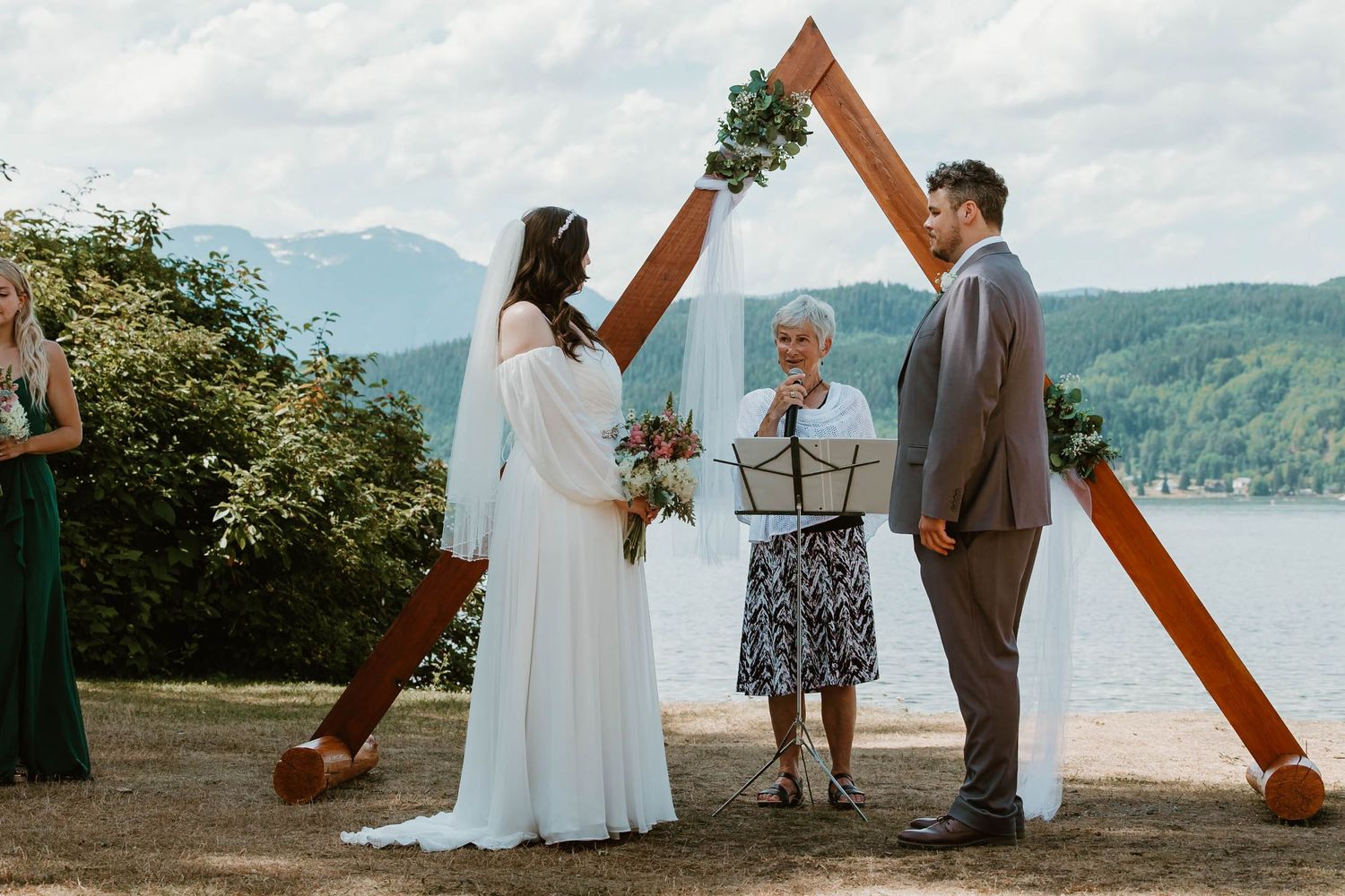 A wedding ceremony takes place beneath a wooden triangle arch with mountain and lake views.