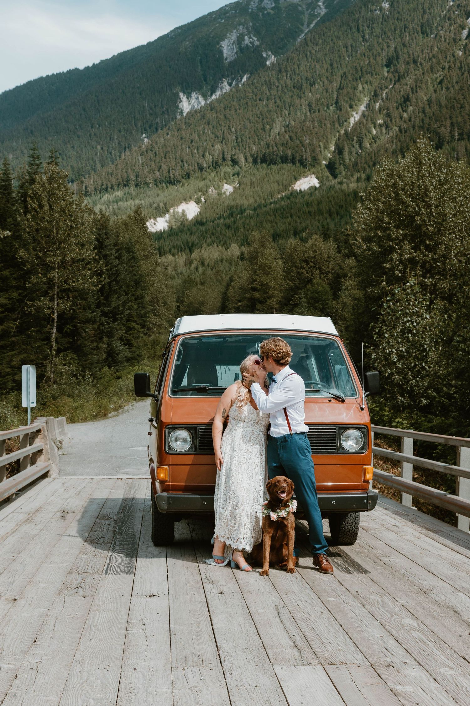 Vintage orange van parked on wooden bridge with mountain backdrop and evergreen forest scenery.
