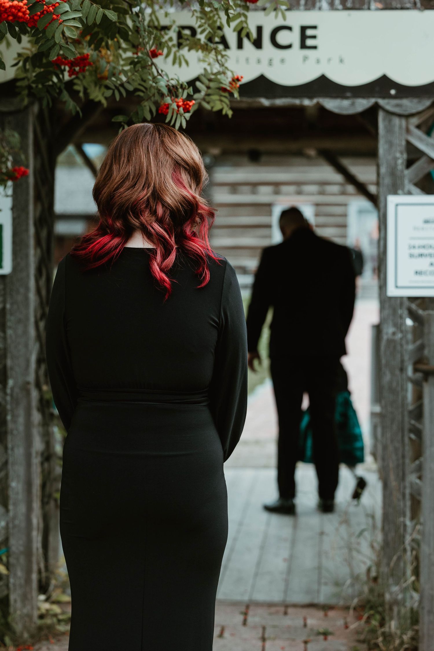 Person with burgundy hair wearing black dress looking towards entrance sign.