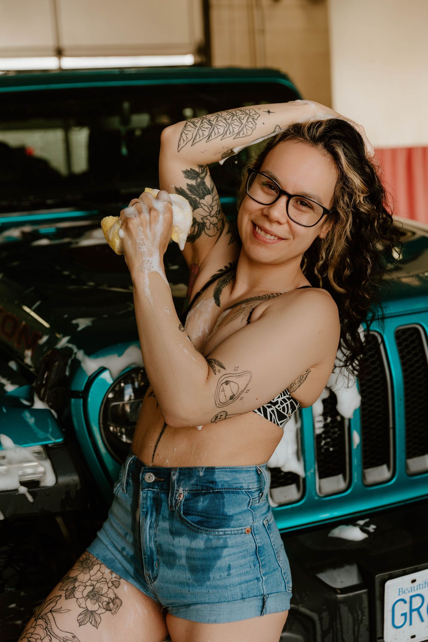 Artistic shot of person in denim shorts washing teal Jeep with visible tattoos and glasses.