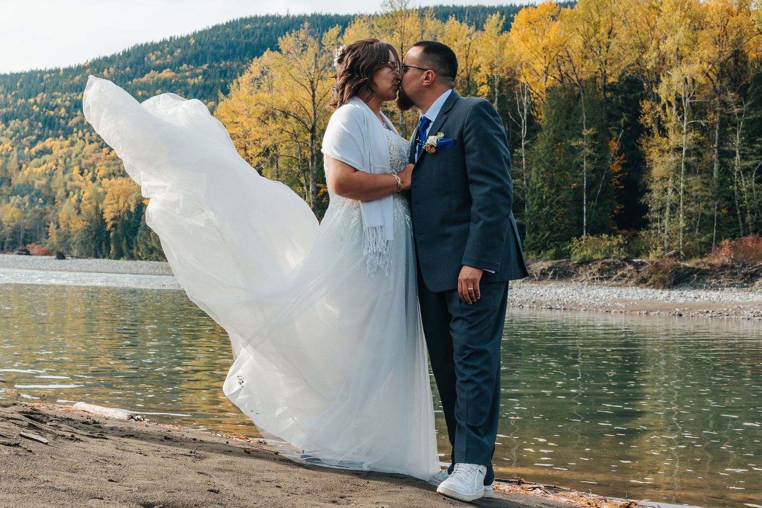 Wedding couple embraces on beach with flowing white dress against autumn trees and lake.