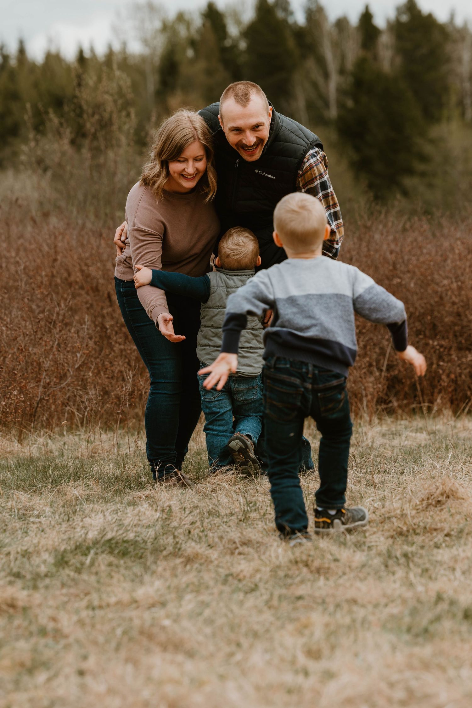 Family playing and running together in a field with pine trees in background.