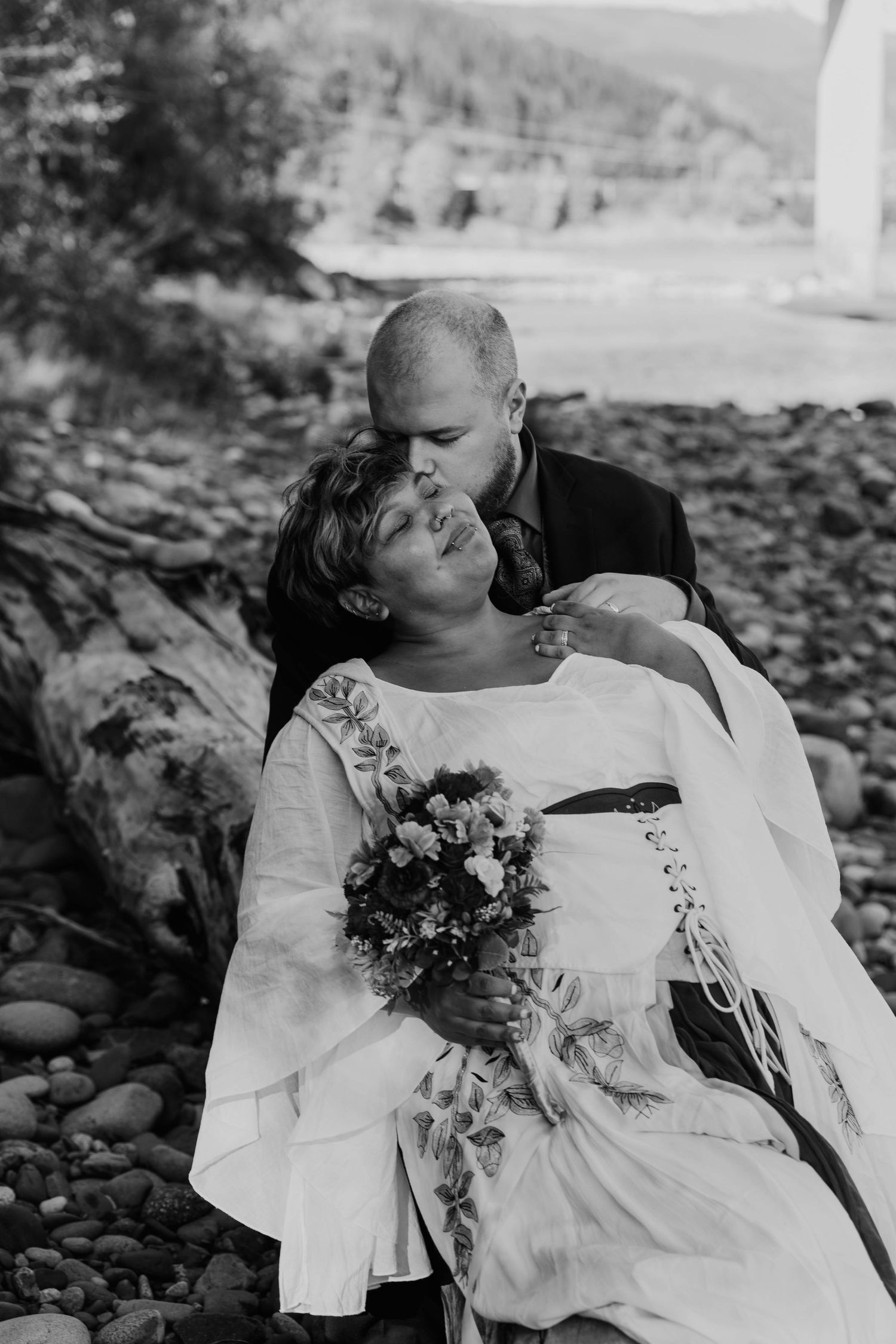 Black and white photo of newlyweds sharing tender moment on rocky shoreline.