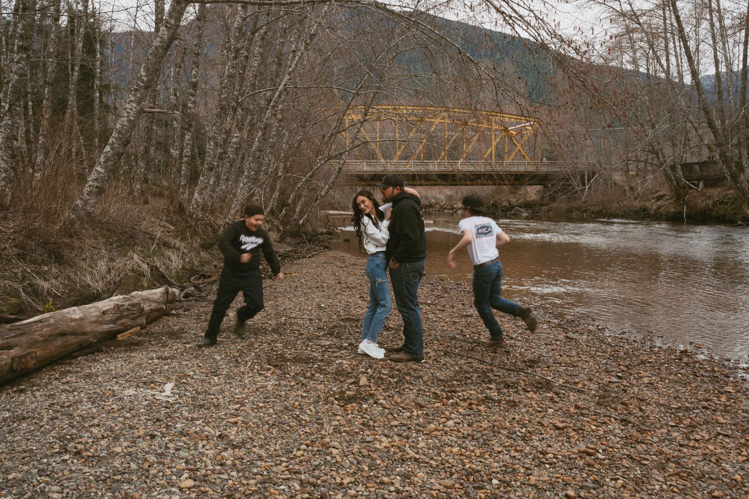 Group enjoying a playful moment by a river with a rustic bridge in the background on a cloudy winter day.