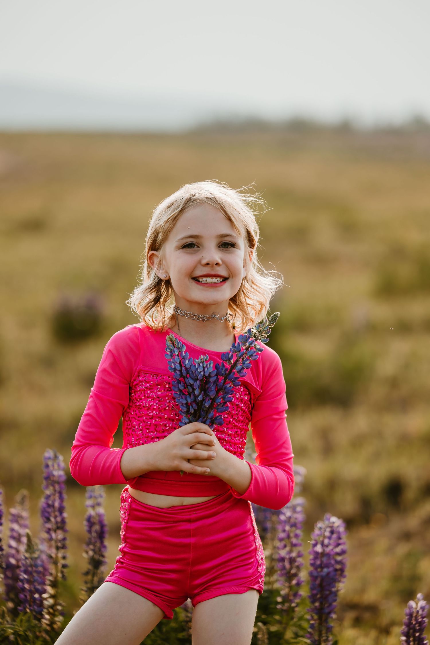 A child in bright pink outfit holds freshly picked purple lupine flowers while standing in a meadow at sunset.