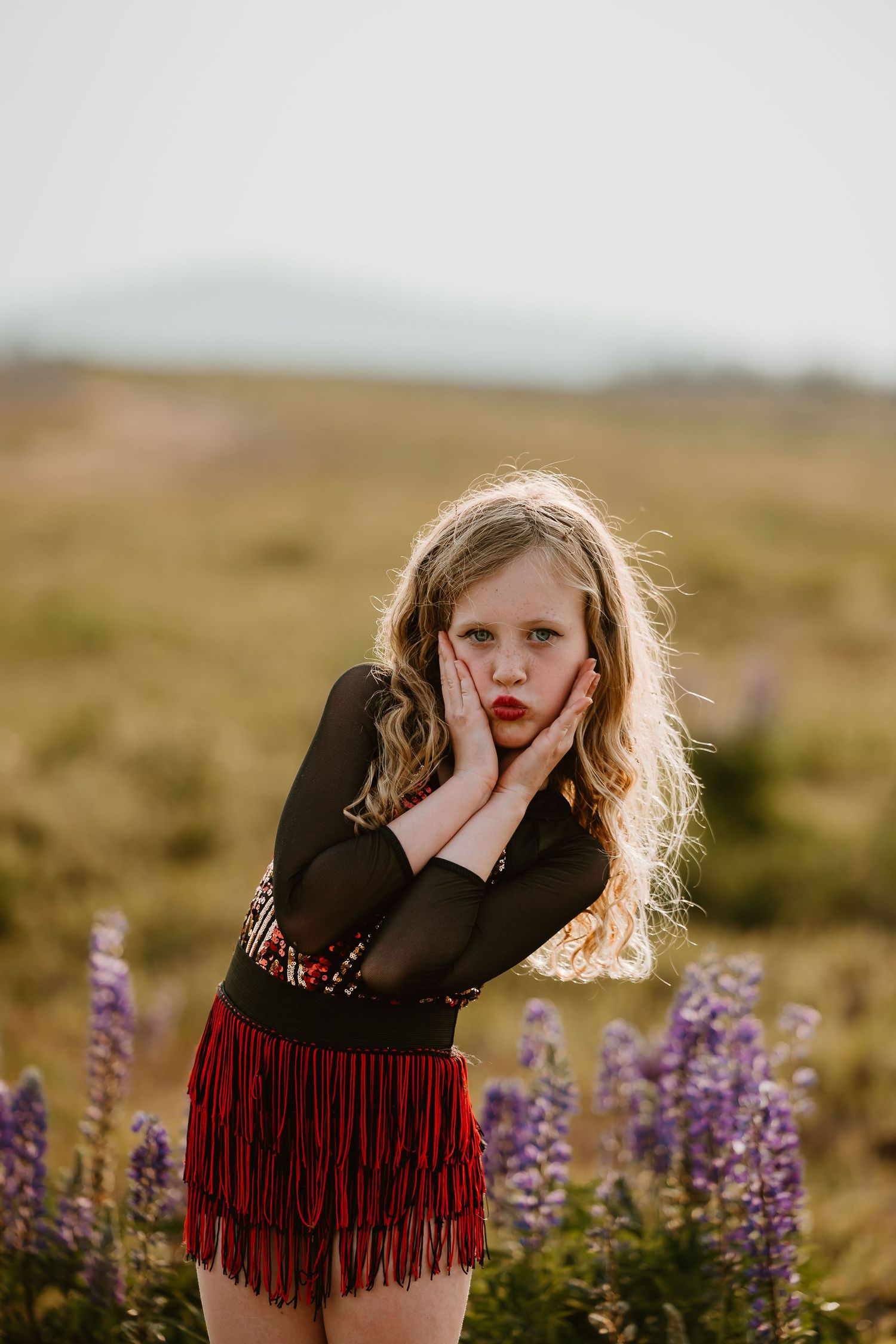A young photographer stands in a wildflower meadow at sunset, wearing a black top with red fringe and making an expressive pose.