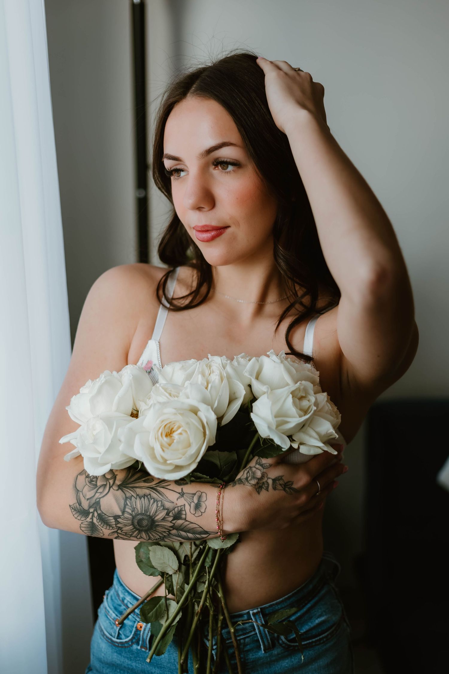 A woman in a strapless top holds white roses against a light window backdrop in a romantic boudoir style photo.