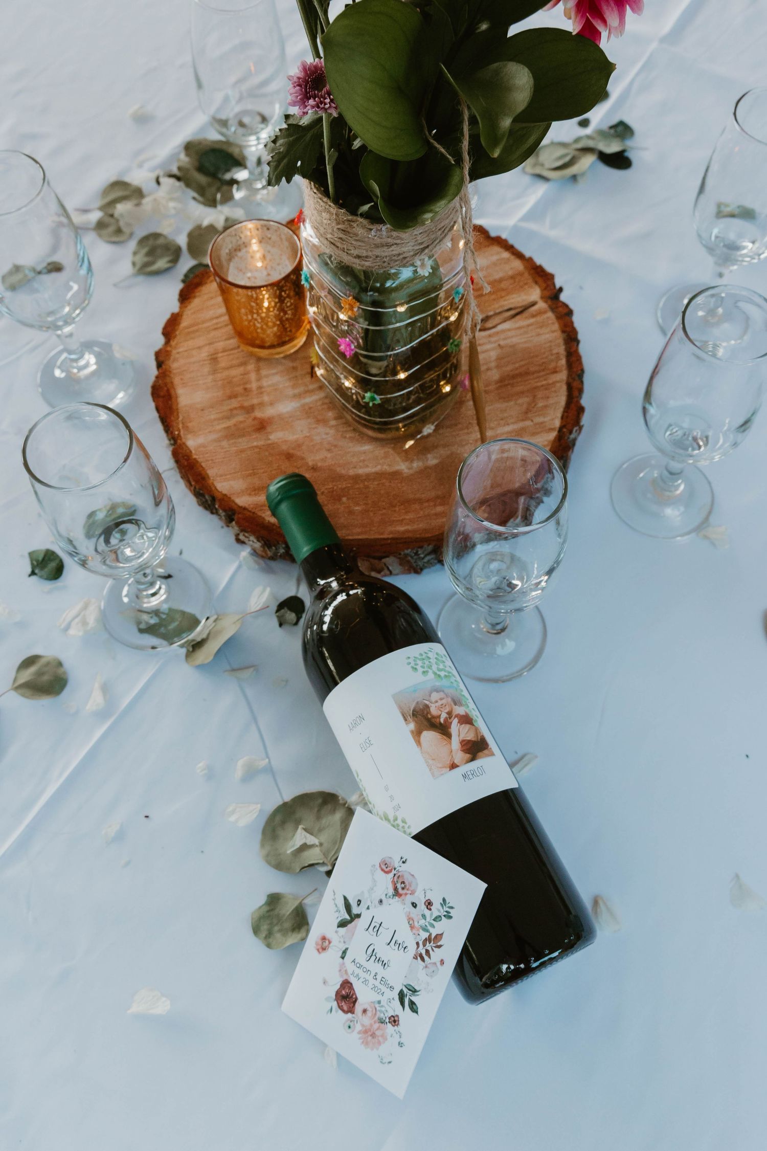 Wedding table centerpiece with wine bottle, glasses and wooden slice on white tablecloth.