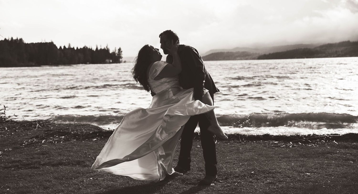 A romantic black and white photo of a couple embracing by the ocean shore with flowing dress fabric.