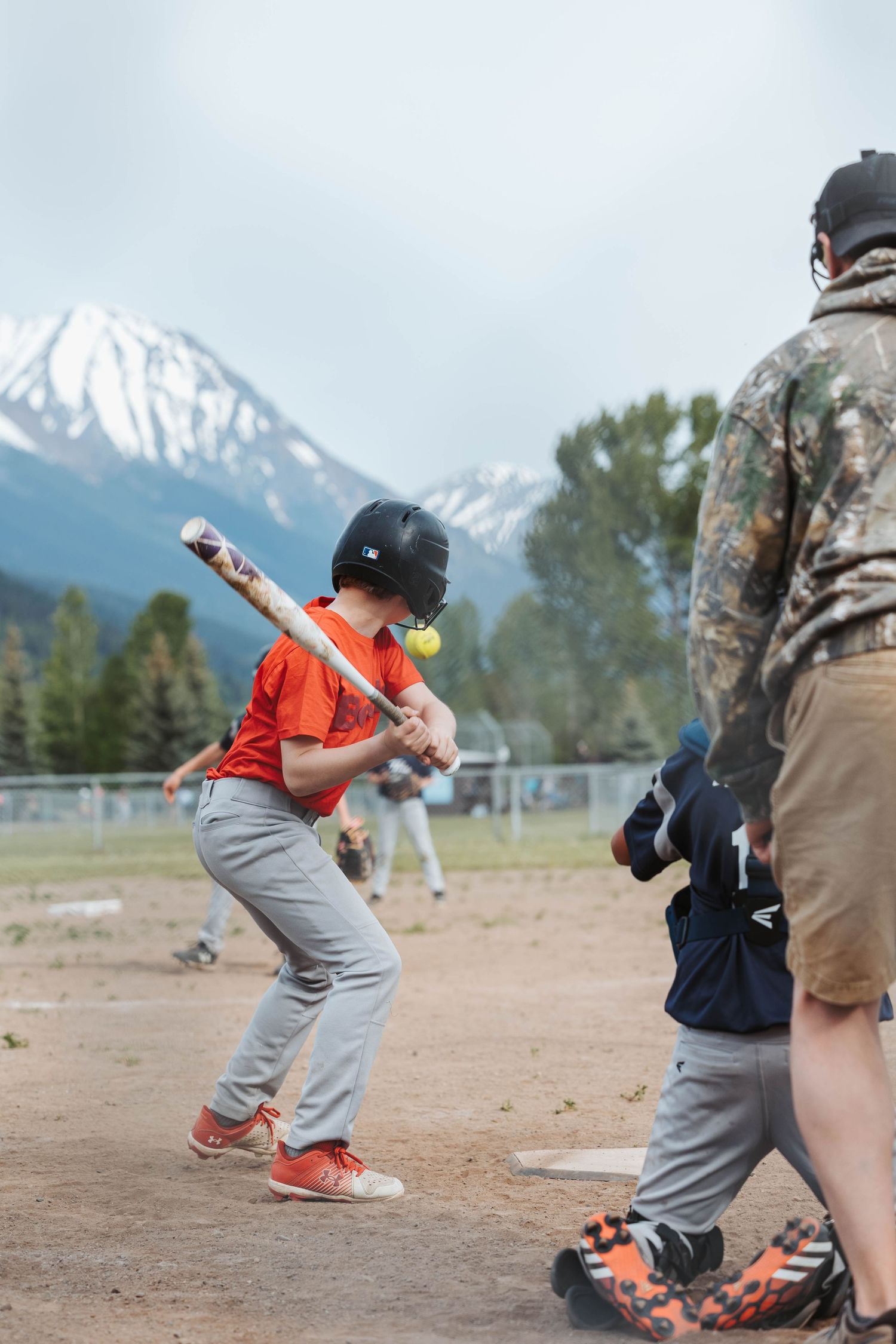 Baseball batter in orange and gray uniform prepares to swing with snow-capped mountains visible.