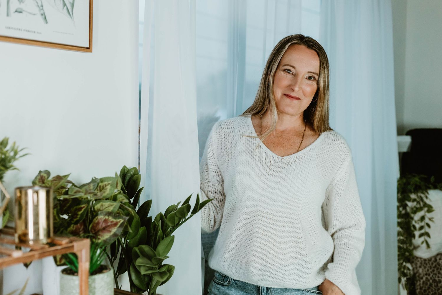 Casual indoor portrait by window with plants wearing white sweater in bright natural light.