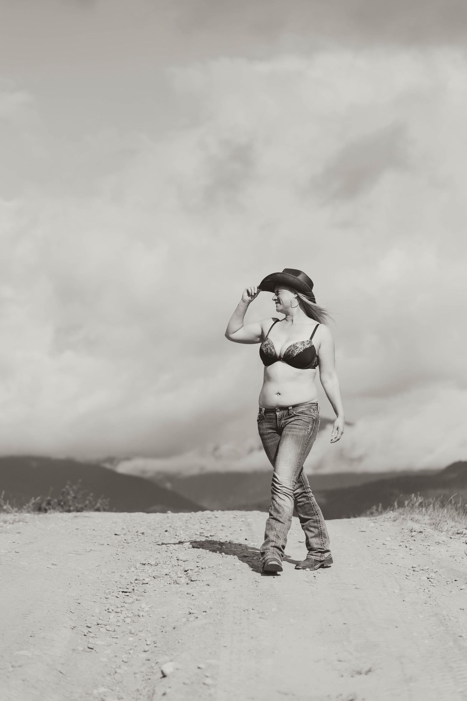 Black and white portrait of figure walking on dirt path against cloudy mountain backdrop.
