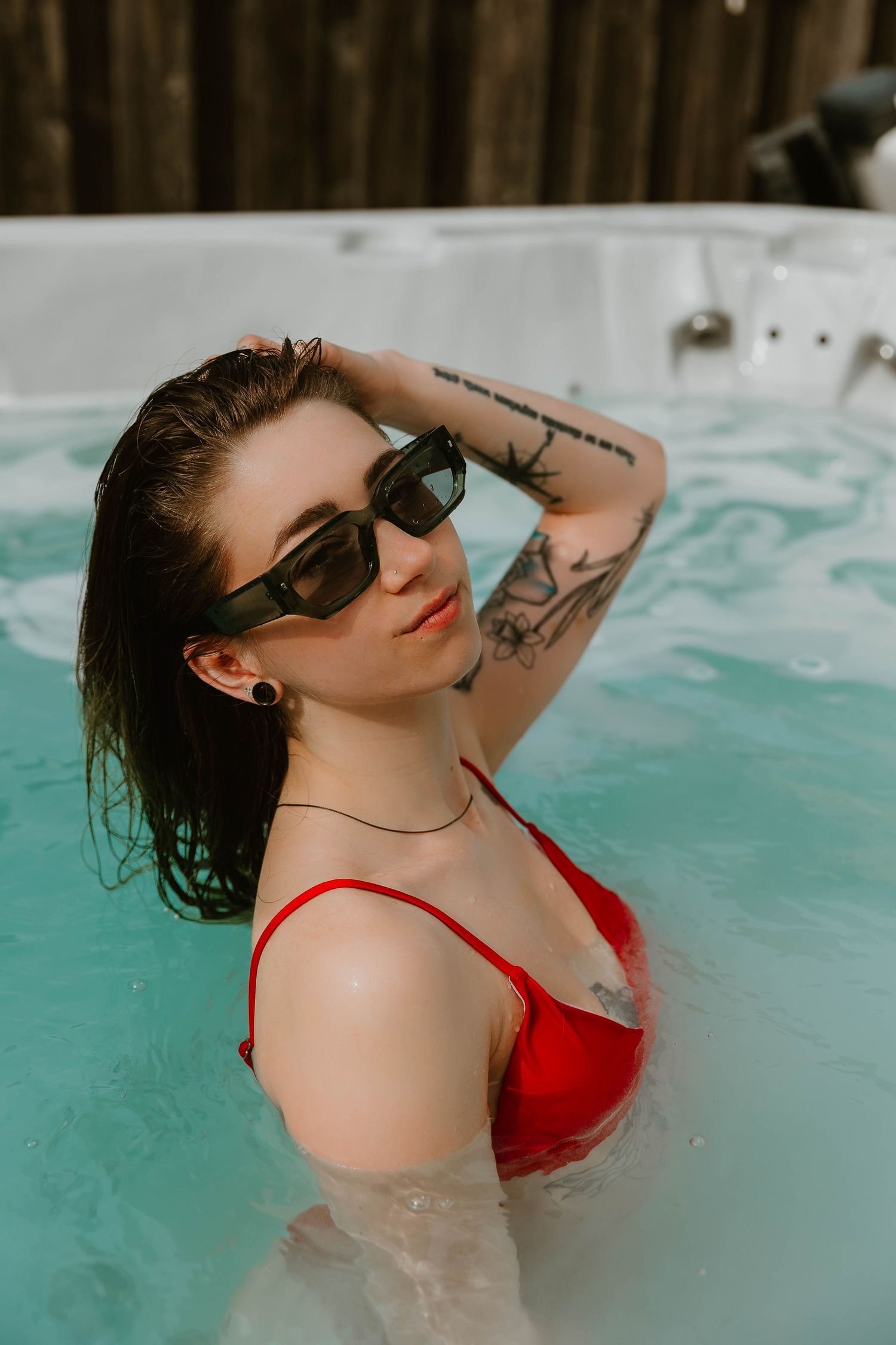 A person wearing sunglasses and a red swimsuit poses in turquoise water with visible tattoos and wet dark hair.