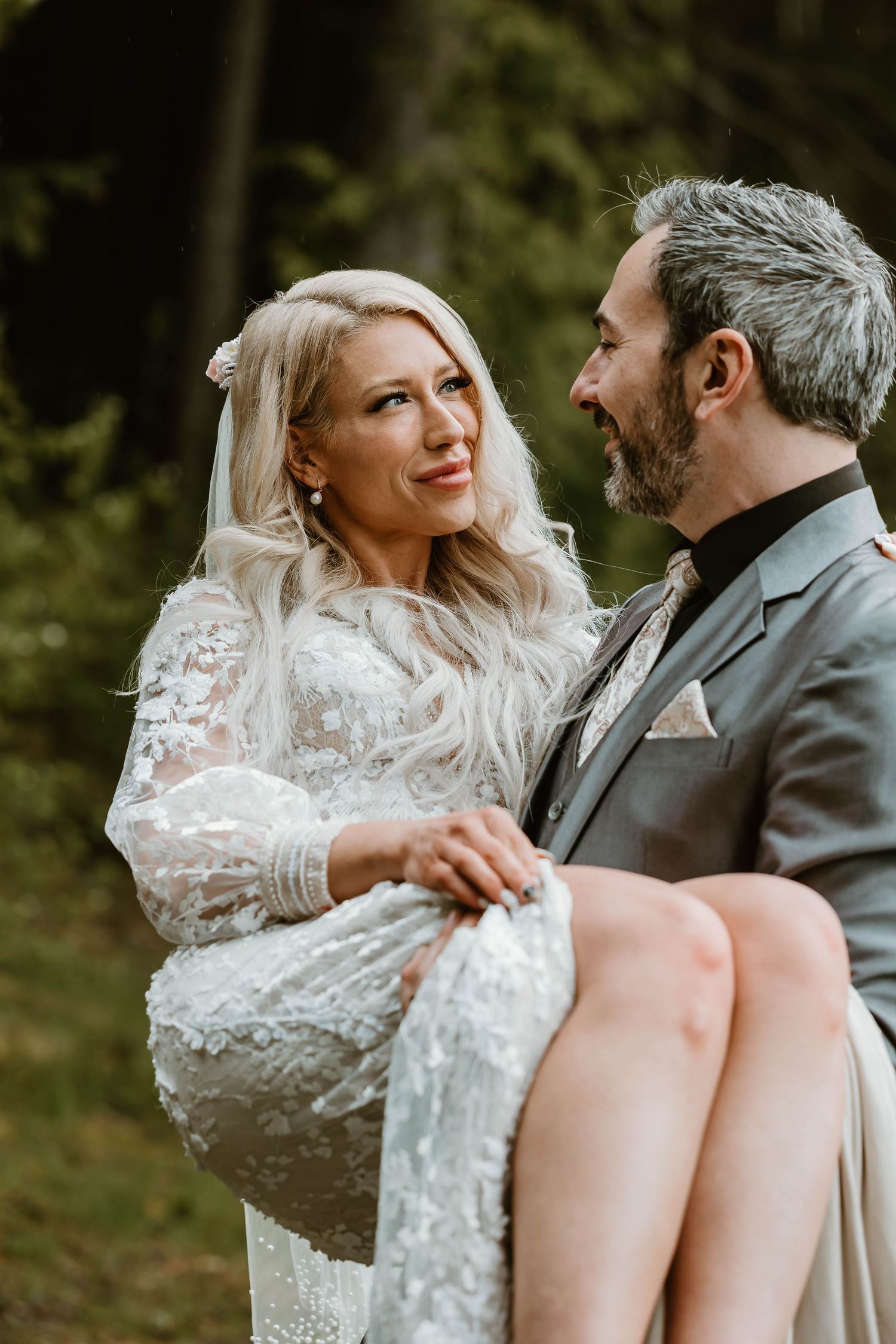 A romantic wedding couple embraces in the woods, with the bride wearing a lace gown and veil.