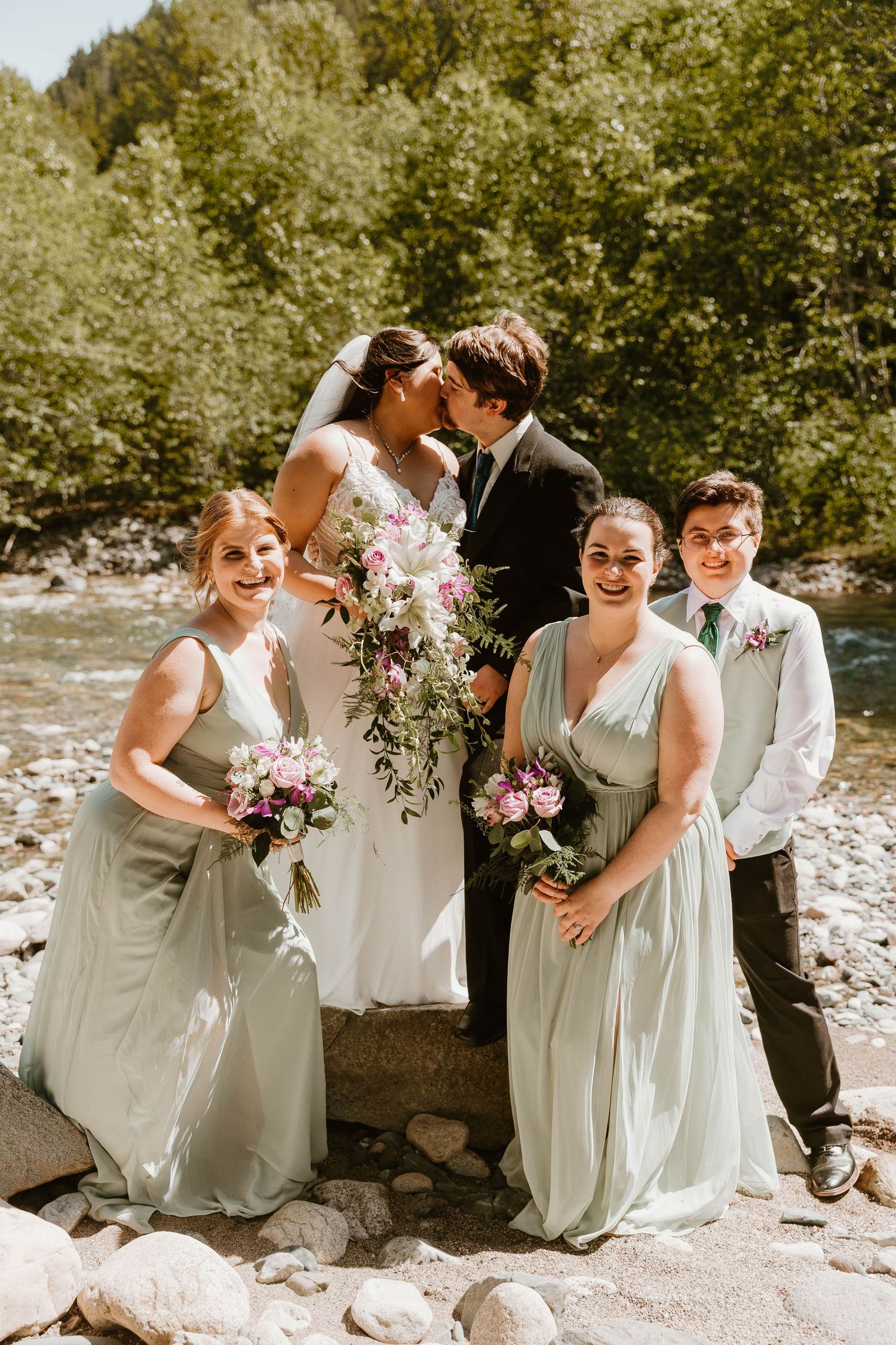 Wedding party poses together on rocky riverbank in forest setting with sage green bridesmaid dresses and floral bouquets.