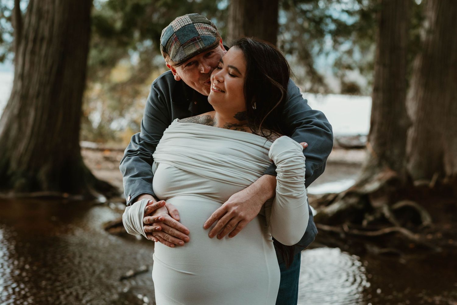 A couple shares an intimate moment in a forest setting during a maternity photoshoot.
